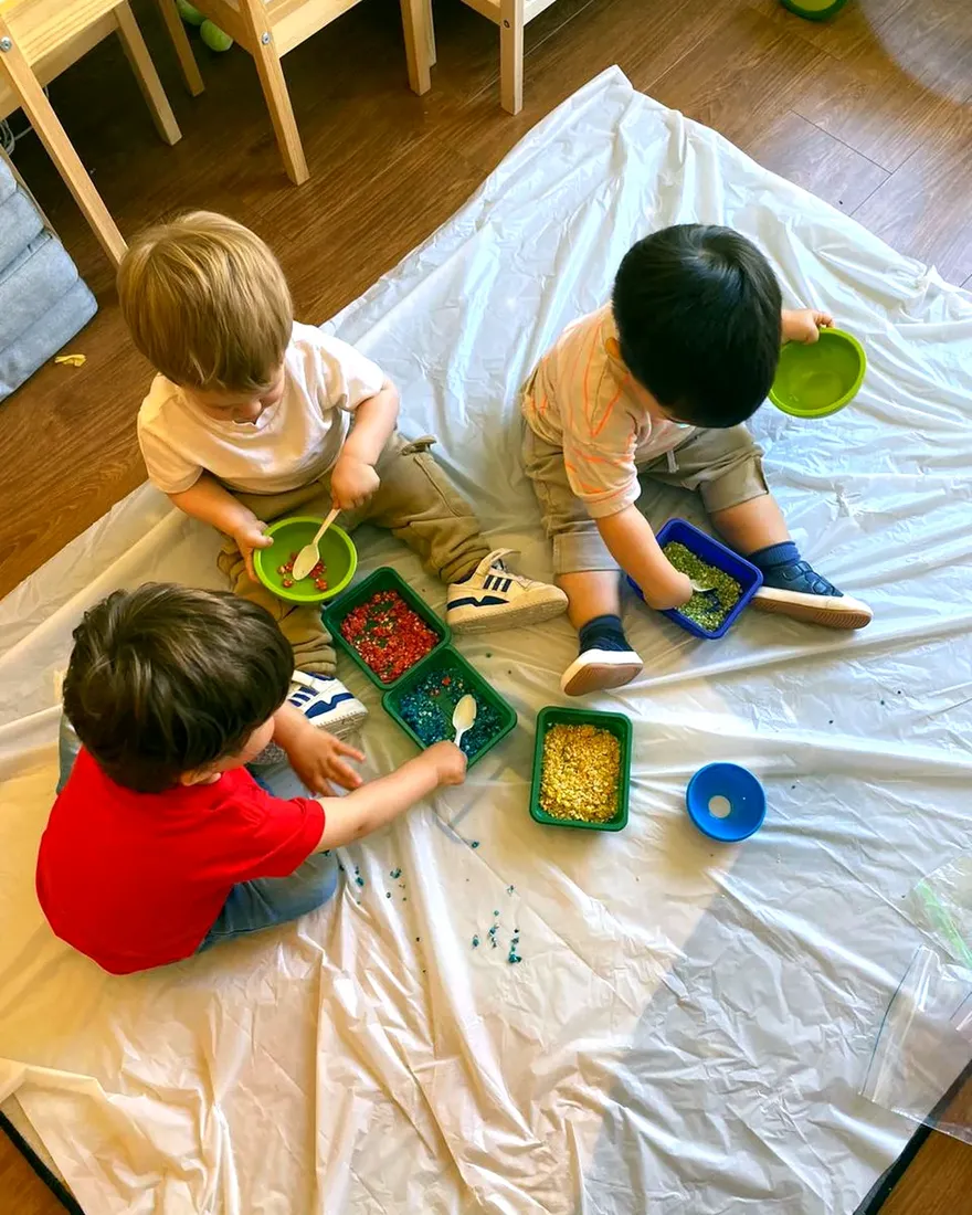 Tres niños juegan con arroz de colores en bandejas, sentados sobre un plástico.