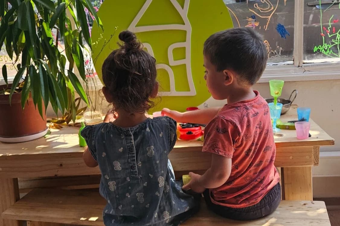 Dos niños sentados a una mesa, junto al isotipo de La Casita, frente a una ventana con dibujos.
